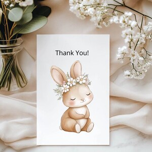 K&ouml;nnte beinhalten: Eine wei&szlig;e Karte mit dem Text "Thank You!" zeigt eine Aquarellillustration eines sitzenden Hasen mit einem Blumenkranz. Der Hase ist hellbraun mit rosa Akzenten, und die Blumen sind wei&szlig;. Die Karte ist von wei&szlig;en Blumen und Gr&uuml;n umgeben.