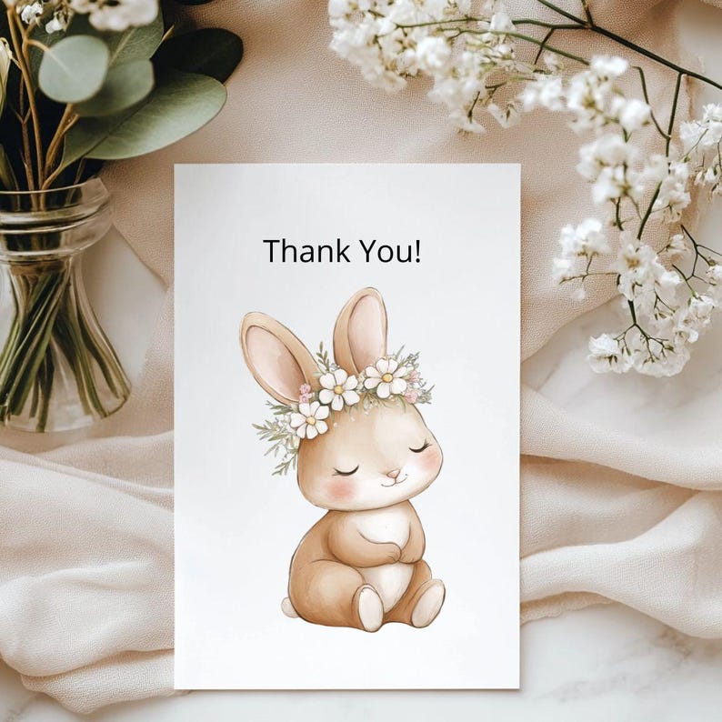 K&ouml;nnte beinhalten: Eine wei&szlig;e Karte mit dem Text "Thank You!" zeigt eine Aquarellillustration eines sitzenden Hasen mit einem Blumenkranz. Der Hase ist hellbraun mit rosa Akzenten, und die Blumen sind wei&szlig;. Die Karte ist von wei&szlig;en Blumen und Gr&uuml;n umgeben.