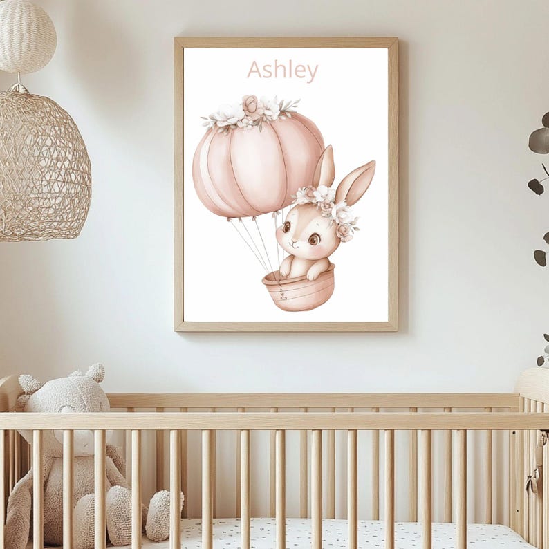 May include: A framed print with a watercolour illustration of a bunny in a hot air balloon. The balloon is a soft pink, decorated with flowers, and the bunny wears a floral crown. The name "Ashley" is written at the top. The artwork is in a light wood frame.