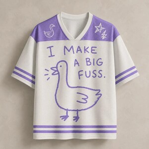 May include: A white and purple V-neck jersey with short sleeves and a mesh texture. The front features a purple line drawing of a goose, a star, a lightning bolt, and the text I MAKE A BIG FUSS. Two purple stripes accent the sleeves and hem.