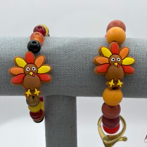 May include: Two beaded bracelets with a Thanksgiving theme. Each bracelet features a cartoon turkey charm with yellow, orange, and red feathers. The bracelets have a mix of colorful beads in shades of brown, orange, and yellow.