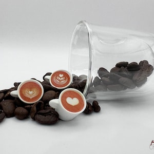 Könnte beinhalten: Drei Miniatur-Kaffeetassen aus weißer Keramik mit Latte-Art-Designs, umgeben von Kaffeebohnen und einer durchsichtigen Glas Tasse mit dem Wort "lavito" an der Seite.