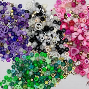 Könnte beinhalten: Eine große Auswahl an bunten Plastikperlen in Lila, Rosa, Grün, Weiß und Schwarz. Die Perlen haben die Form von Sternen, Herzen, Friedenssymbolen und anderen Designs. Die Perlen sind perfekt für die Herstellung von Schmuck, Kunsthandwerk und anderen Projekten.