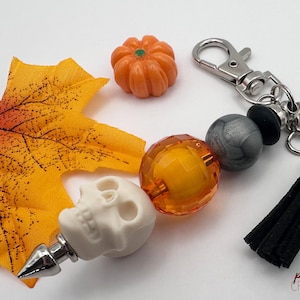 May include: A Halloween-themed keychain with a white skull bottle stopper, orange pumpkin charm, and a black tassel. The keychain also features a clear orange bead and a gray bead. A fall leaf is also present.