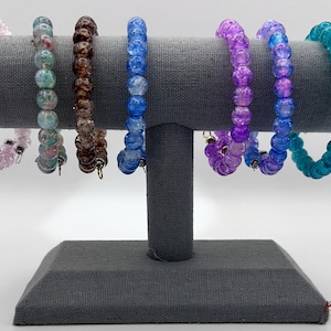 May include: Seven beaded bracelets displayed on a gray stand. The bracelets are made of clear glass beads with different colors and patterns. The colors include pink, teal, brown, blue, and purple.