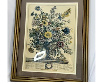 Puede incluir: Lámina floral enmarcada con un vibrante ramo de diversas flores en un jarrón decorativo. La obra de arte incluye una variedad de colores, como amarillo, azul y rojo, sobre un fondo crema. El marco es dorado.