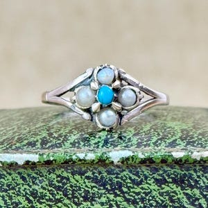 May include: A silver ring featuring a central turquoise stone surrounded by four pearls and four smaller opalescent stones. The ring has an openwork design and is set on a green and black patterned surface.