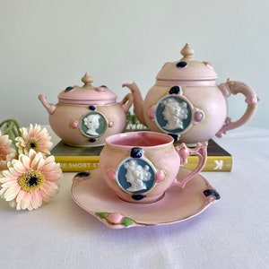 Può includere: Un set da tè rosa con teiera, zuccheriera, tazza e piattino. Il set è decorato con un cammeo bianco del viso di una donna e dettagli rosa e blu.