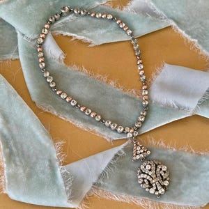 May include: A silver necklace with clear and rose-coloured stones. The necklace has a pendant with a triangular top and a round, floral-like bottom. It is displayed on a light blue ribbon against a tan background.