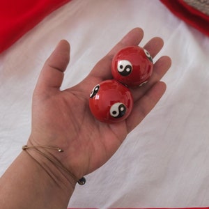 May include: Two red Chinese health balls, each with a black and white yin-yang symbol, are held in an open hand. The balls have a glossy finish, and the yin-yang symbols are distinct. The background is a white fabric.