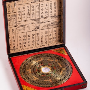 May include: A red wooden box with a hinged lid, open to reveal a compass with a black and gold face. The compass is surrounded by a red felt lining. The inside of the lid is covered in a printed paper with Chinese characters.