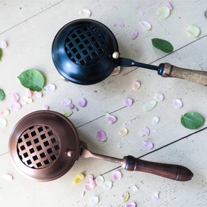 Pode incluir: Dois suportes de flores de metal com cabos de madeira. Um é preto, o outro é cor de cobre. Ambos têm um design redondo em forma de grade na parte superior. Os suportes estão em uma superfície de madeira clara, cercados por pétalas de flores e folhas verdes.