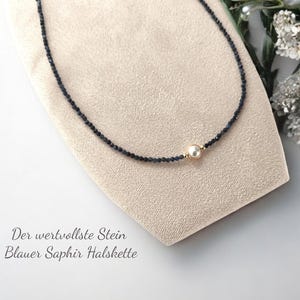 Könnte beinhalten: Eine zarte Halskette aus kleinen schwarzen Saphirperlen mit einem einzigen weißen Perlenakzent. Die Halskette wird auf einer beigefarbenen Stoffoberfläche präsentiert. Der Text "Der wertvollste Stein Blauer Saphir Halskette" ist unterhalb der Halskette sichtbar.