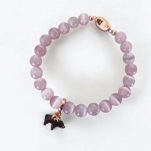 May include: A lilac beaded bracelet with a rose gold clasp and charm. The bracelet features round, polished beads and a small, black charm with a rose gold accent. The bracelet is displayed on a white background.