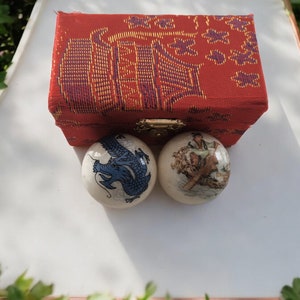 Peut inclure: Deux boules en céramique blanche avec des motifs peints. Une boule représente un dragon bleu, l'autre une scène avec des personnes en vêtements traditionnels. Les boules sont dans une boîte rouge avec un fermoir doré.