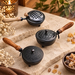 Puede incluir: Tres quemadores de incienso de metal negro con mangos de madera y patas pequeñas. Cada quemador presenta un diseño de tapa distinto: uno con patrón de rejilla, otro con patrón floral geométrico y otro con patrón de puntos perforados. Son redondos.