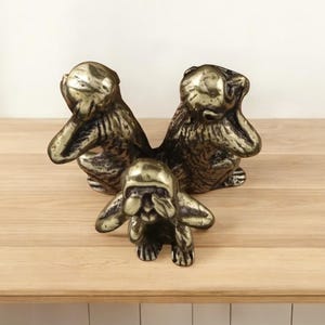 Puede incluir: Escultura de color bronce que representa a los tres monos sabios: No ver el mal, no oír el mal, no hablar el mal. Los monos están sentados, con las manos cubriendo los ojos, las orejas y la boca. La escultura está sobre una superficie de madera.