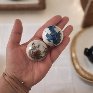 Może przedstawiać: Dwie białe ceramiczne kule z malowanymi wzorami. Jedna kula przedstawia niebieskiego smoka, druga osobę jeżdżącą konno.