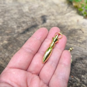 Può includere: Un pendolo dorato con una punta appuntita e un anello nella parte superiore. Il pendolo è tenuto in una mano.