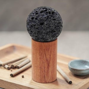 May include: A dark gray, porous sphere sits atop a cylindrical wooden base. The base is on a wooden tray, along with several small, rolled items and a small, light blue bowl. The items appear to be for aromatherapy or a similar purpose.