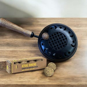 Pode incluir: Queimador de incenso preto com cabo de madeira e recortes decorativos de luas e estrelas. Uma pequena caixa com o texto "ATRAE DINERO" e duas bolas de incenso redondas estão ao lado do queimador. O queimador e a caixa estão sobre uma superfície de madeira.