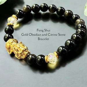 Könnte beinhalten: Ein Feng Shui Armband aus Gold, Obsidian und Citrin. Das Armband besteht aus schwarzen, runden Obsidianperlen, die mit Citrinsteinen und goldenen Akzenten durchsetzt sind. Ein goldfarbener Pixiu-Anhänger ist ein herausragendes Merkmal.