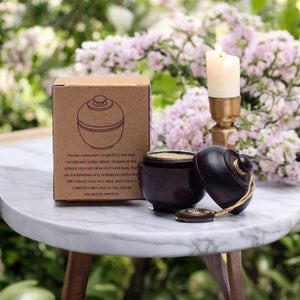 May include: A wooden jar with a lid, handcrafted from Indian wood. The jar is brown with a natural wood grain and has a rope handle. The jar is sitting on a white marble table with a candle and flowers in the background. The jars have been made from the best handpicked Indian Wood. Shaped by the artisans on small lathe and hand tools, they are smoothened and polished to perfection. With natural colour and veins, each jar is a collector's item and no two pieces are exactly identical.