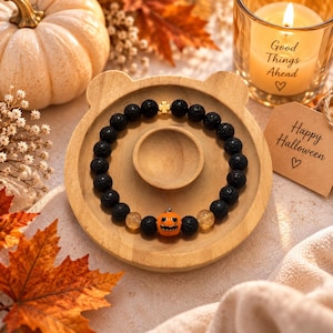 Può includere: Un braccialetto di perline con pietre laviche nere, due perline gialle traslucide e una perlina centrale arancione a forma di zucca. Un piccolo ciondolo dorato a forma di quadrifoglio è posizionato nella parte superiore della composizione.