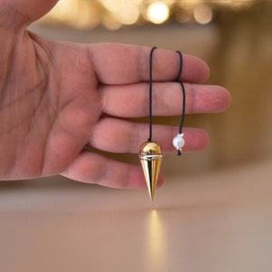 Könnte beinhalten: Ein goldfarbenes Pendel mit einer schwarzen Schnur und einer weißen Perle. Das Pendel hat die Form eines Kegels mit einer abgerundeten Spitze.