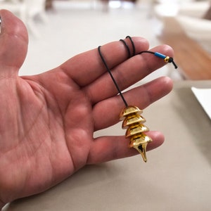 Puede incluir: Un péndulo de color dorado con una punta puntiaguda y una serie de discos redondeados apilados. El péndulo está unido a un cordón negro con una cuenta azul.