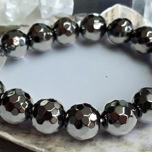 Könnte beinhalten: Ein silberfarbenes Armband mit facettierten, kugelförmigen Perlen. Die Perlen haben einen metallischen Glanz und sind aufgereiht. Das Armband wird auf einer weißen Oberfläche präsentiert. Die Perlen haben einen Durchmesser von ca. 1,5 cm.