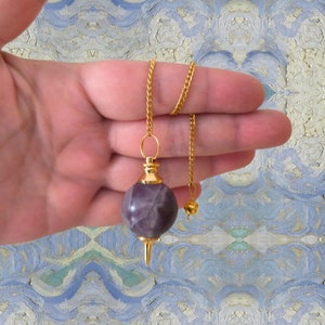 Op de afbeelding: Een gouden ketting met een paarse amethist kristallen slinger. De slinger is rond en heeft een puntige punt. De ketting wordt in een hand gehouden.