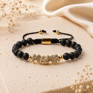 May include: A black lava bead bracelet featuring a central gold-toned charm that spells out the word MAMA in sparkling crystals. The bracelet includes a black cord with an adjustable sliding knot closure and two small gold-toned end beads.