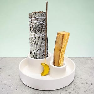 Könnte beinhalten: Ein weißer Keramik-Räucherhalter mit einem Salbeibündel, einem Palo Santo-Stab und einem Räucherstäbchen. Der Halter hat einen gelben Halbmond-Anhänger. Der Salbei ist mit weißem Faden umwickelt.