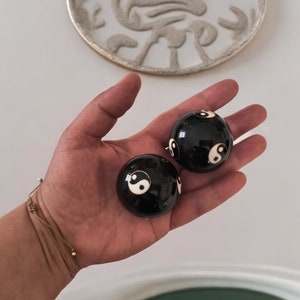 Op de afbeelding: Twee zwarte en witte yin yang ballen in een hand. De ballen zijn ongeveer 5 cm in diameter en hebben een gouden rand rond het yin yang symbool.