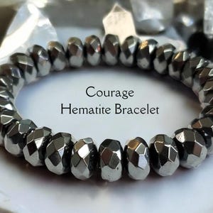 Könnte beinhalten: Ein Armband aus facettierten, metallisch grauen Hämatitperlen. Das kreisförmige Armband liegt auf einer weißen Oberfläche. Der Text auf dem Bild lautet "Courage Hematite Bracelet".