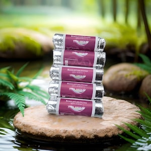 Könnte beinhalten: Fünf folienverpackte Packungen KOHLETABLETTEN, jede mit einem lila Etikett und dem Text "HOLA SMOKE". Die Packungen sind auf einem Stein in einem Teich gestapelt, mit grünen Pflanzen im Hintergrund.