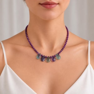 Op de afbeelding: Een ketting met een streng kleine, ronde, dieppaarse kralen. Het midden van de ketting is versierd met vijf druppelvormige hangers in verschillende tinten doorschijnend groen, blauw en paars, gerangschikt in een symmetrisch patroon.