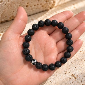 Pode incluir: Uma pulseira elástica composta por contas redondas e porosas de pedra de lava preta. O design apresenta uma única conta de metal prateado em forma de bola de futebol com detalhes pentagonais pretos e prateados, integrada no fio de contas.