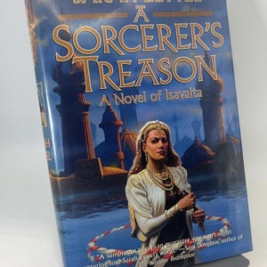 May include: A hardcover book titled "Sorcerer's Treason" by Sarah Zettel. The cover features a woman in ornate clothing, a fantasy landscape, and the text "A Novel of Isavalta". The book is displayed on a metal stand.