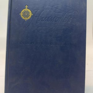 May include: A dark blue hardcover book titled Hammond's Complete World Atlas. The front cover features a gold-colored compass rose emblem and embossed cursive lettering. The book has a vertical spine on the left side and a rectangular shape.