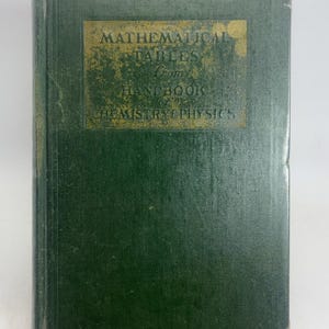 May include: A vintage green hardcover book titled "MATHEMATICAL TABLES From HANDBOOK of CHEMISTRY & PHYSICS." The book is held upright by a gray metal book stand. The cover has a textured appearance with gold lettering.