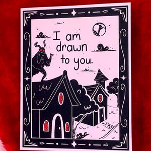 May include: A pink card with a black border and a black and white illustration of a moth with red eyes, two houses, and a moon. The text on the card reads "I am drawn to you."
