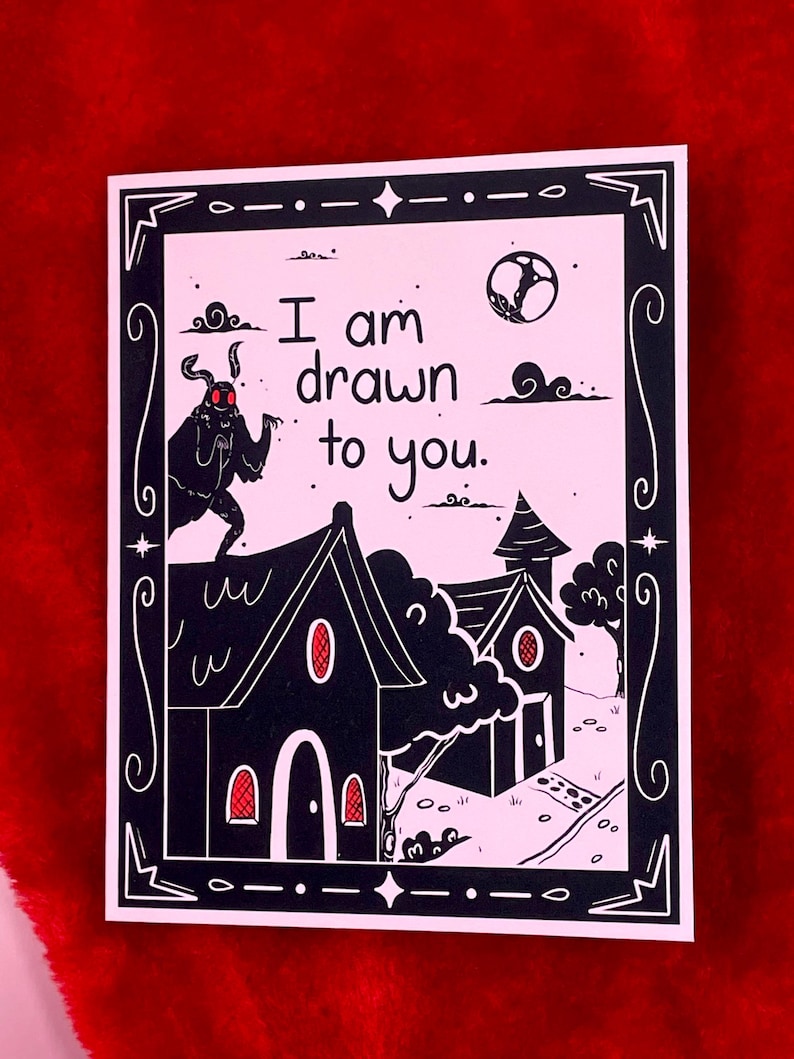 May include: A pink card with a black border and a black and white illustration of a moth with red eyes, two houses, and a moon. The text on the card reads "I am drawn to you."