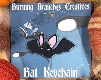 May include: A black bat keychain with pink inner ears and a smiling face, attached to a silver star charm. The keychain is on a blue background with the text "Bat Keychain" and "Burning Branches Creatives".