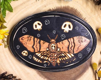 May include: Oval wooden plaque with a black border and a detailed wood-burned design. The central image is a moth with a skull-shaped head, surrounded by small skull motifs. The moth's wings have a striped pattern.