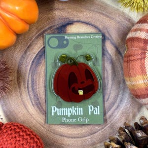 May include: A phone grip featuring a smiling, red pumpkin face with a single tooth, labeled "Pumpkin Pal." The grip is on a green background with the text "Burning Branches Cretive." The image is surrounded by fall-themed decorations.