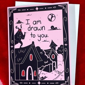 May include: A pink greeting card with a black and white illustration of a moth, two houses, and a moon. The text on the card reads "I am drawn to you."