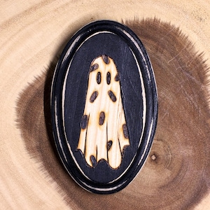 May include: Oval wooden plaque featuring a ghost design. The ghost is light-colored with dark spots, set against a black background within a black and white oval frame. The plaque is placed on a wooden surface.