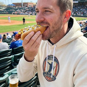 May include: A person eating a hot dog at a baseball game. The hot dog is in a bun and topped with mustard and chopped onions. The person is wearing a cream-colored hoodie with a USA graphic.